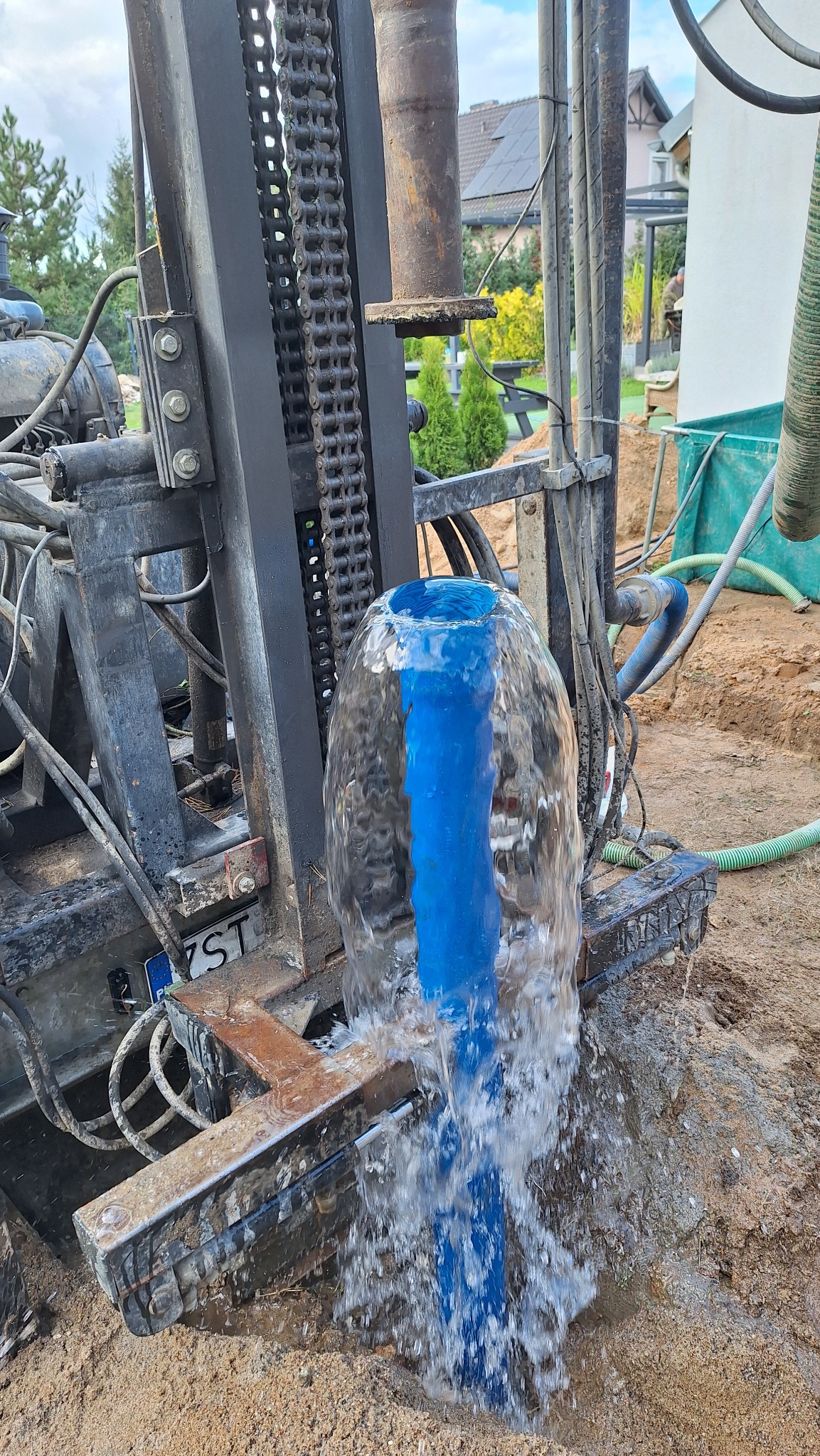 Woda wytryskuje spod maszyny wiertniczej z widoczną tablicą rejestracyjną i fragmentem domu w tle.