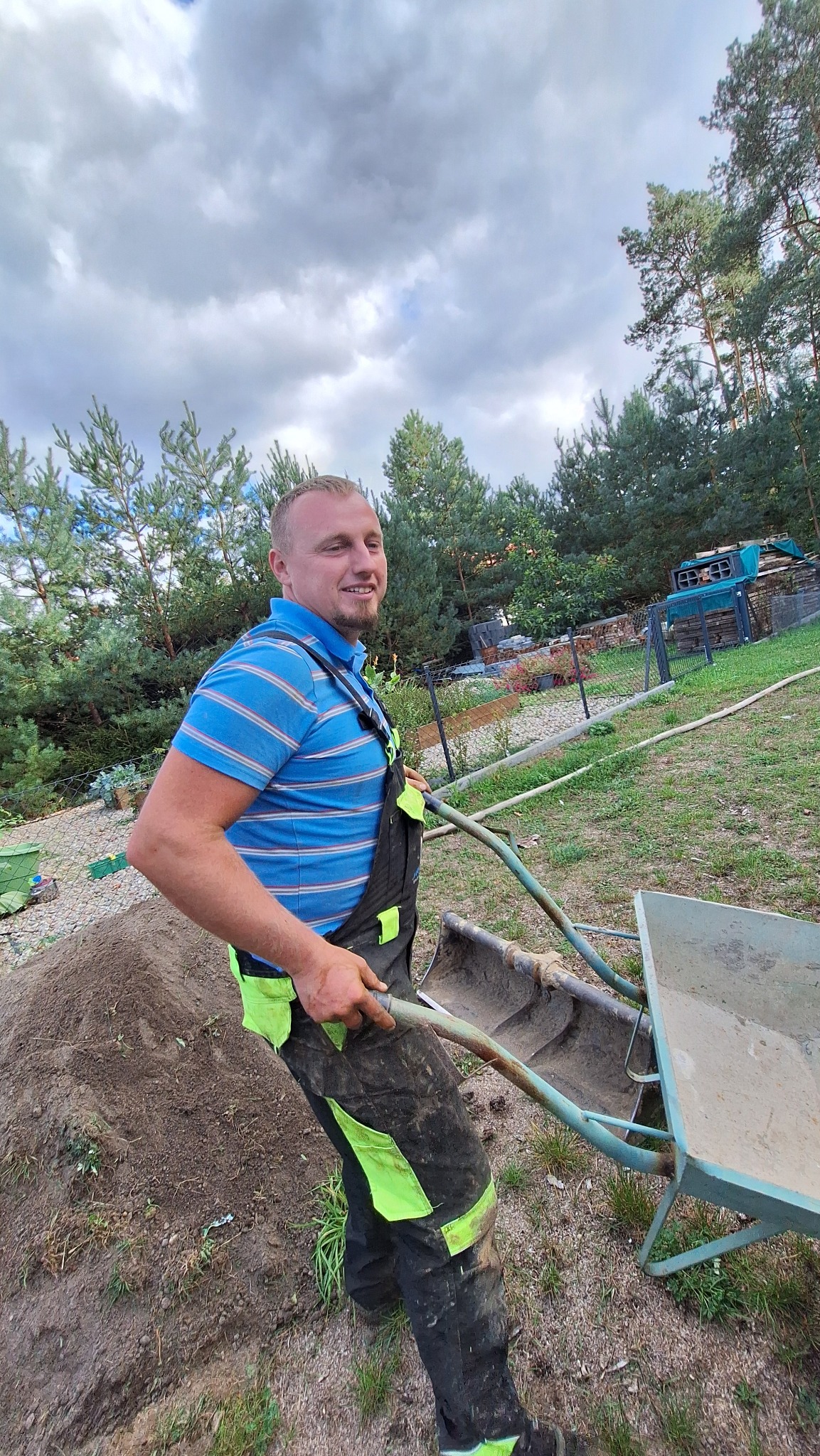 Uśmiechnięty mężczyzna w roboczym kombinezonie z neonowymi wstawkami, pchający brudną taczkę pełną ziemi na tle zielonego ogrodu z drzewami iglastymi i zachmurzonym niebem.