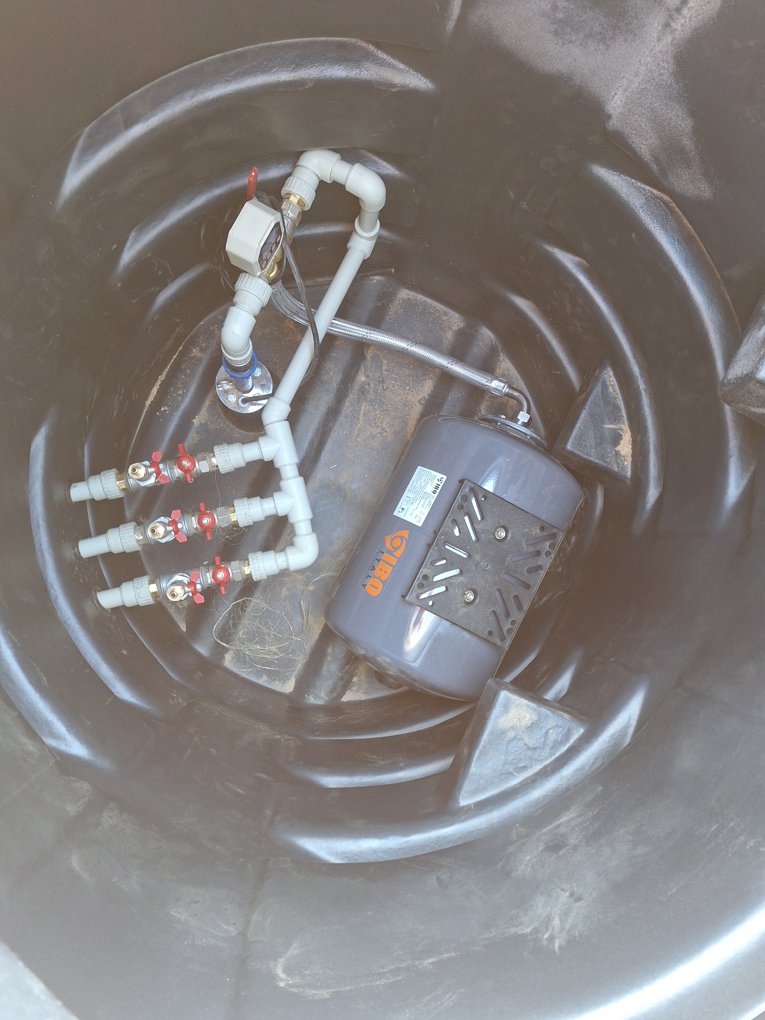 Wnętrze czarnego zbiornika z zainstalowanym szarym zbiornikiem ciśnieniowym, trzema zaworami kulowymi z czerwonymi uchwytami i elementami instalacji wodnej.