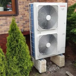Zewnętrzna jednostka pompy ciepła Toshiba ESTIA, zainstalowana na betonowych bloczkach przy ścianie z cegły klinkierowej w Malanowie, otoczona tujami i kamieniami.