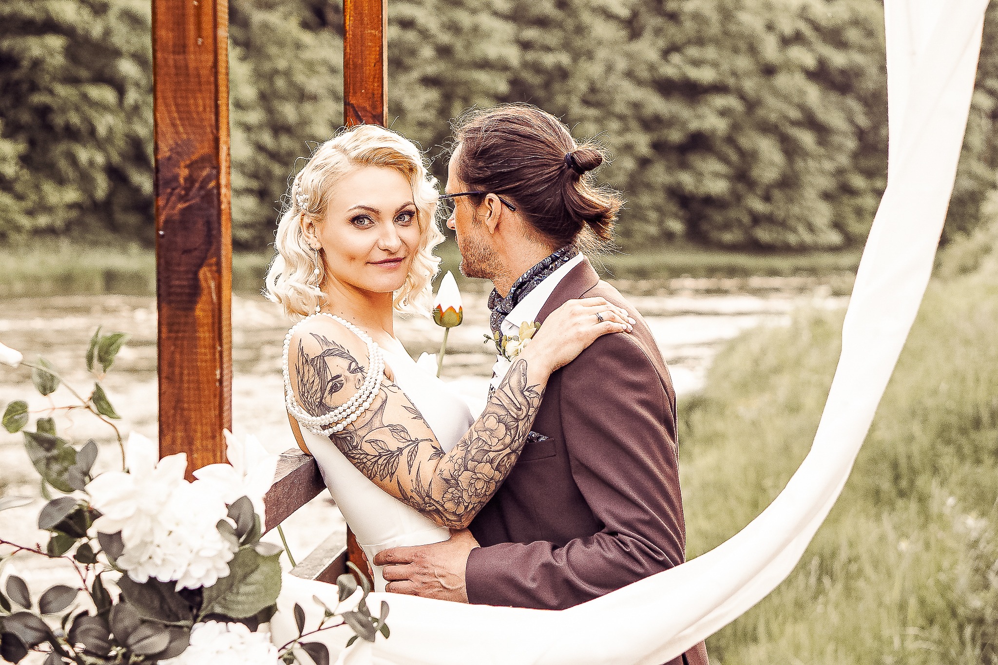 Para młoda: blondwłosa panna młoda z tatuażami na ramionach spogląda w obiektyw, obejmowana przez pana młodego w brązowym garniturze, na tle drewnianej konstrukcji i zieleni.