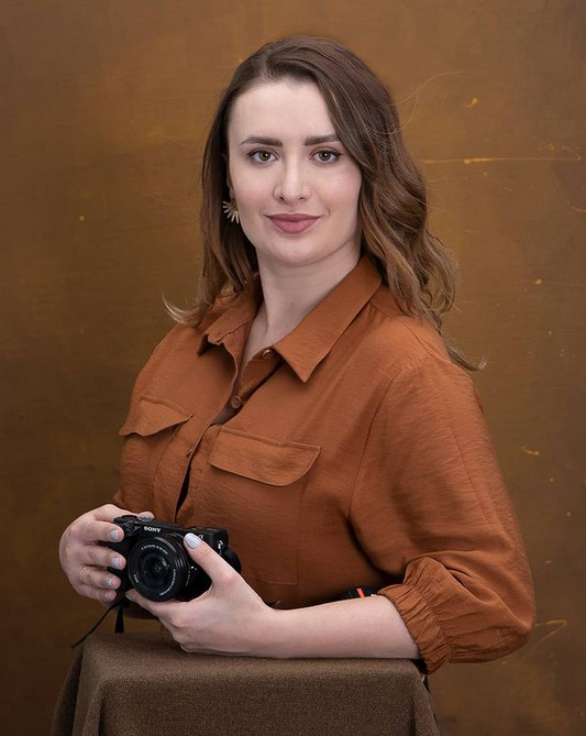 Portret kobiety z aparatem Sony na tle abstrakcyjnej, złoto-brązowej tekstury, ubrana w brązową koszulę z długim rękawem.