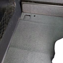 MARTOM Martyna LANGER - Szary dywanik w bagażniku samochodu z siatką po lewej stronie i fragmentem białej tkaniny w prawym dolnym rogu.
