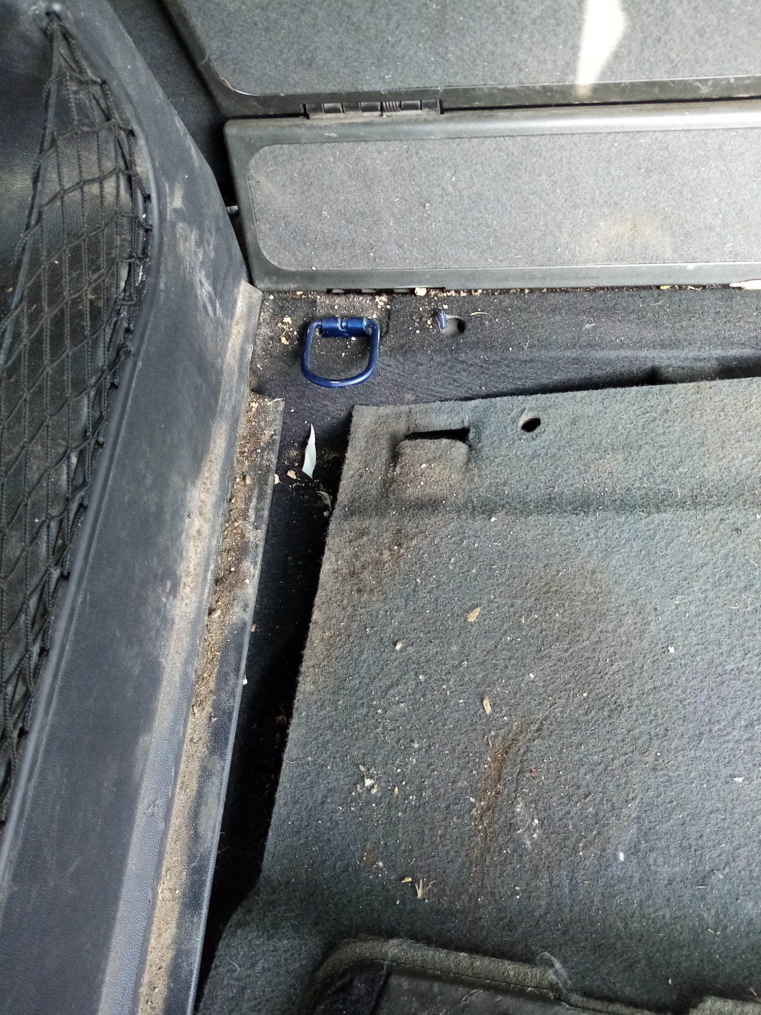 Brudny bagażnik samochodu z widoczną siatką, uchwytem mocującym i dywanikiem pokrytym okruchami i kurzem przed czyszczeniem.