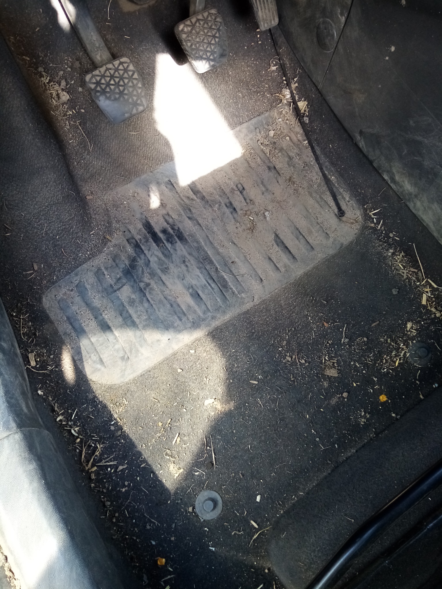 Bardzo brudna podłoga samochodu z gumowym dywanikiem pokrytym piaskiem, błotem i resztkami roślin, widoczne pedały.