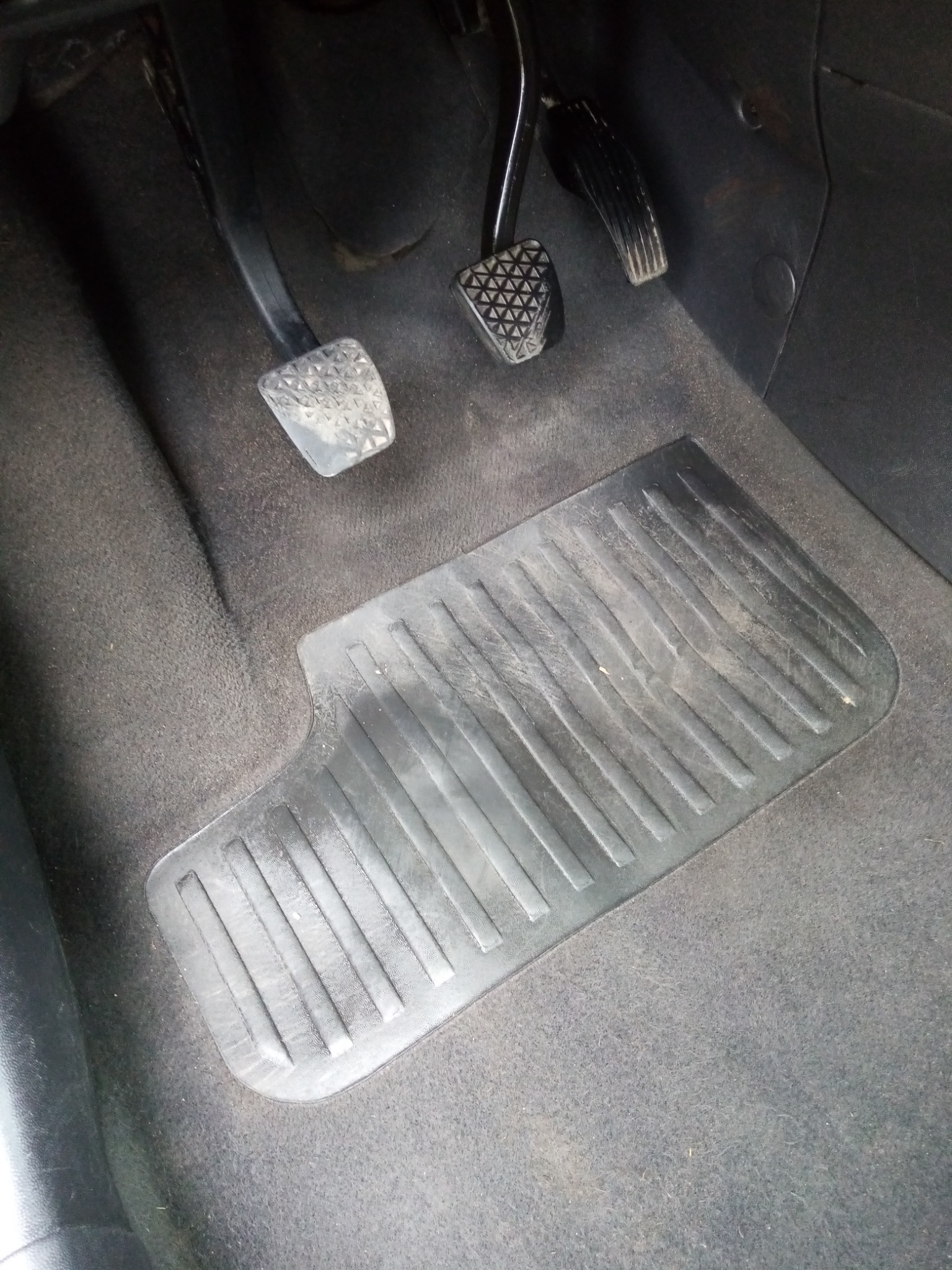 Brudna gumowa wycieraczka samochodowa z podłużnymi rowkami leżąca na ciemnej, lekko zabrudzonej wykładzinie podłogowej w aucie, widoczne pedały gazu, hamulca i sprzęgła.