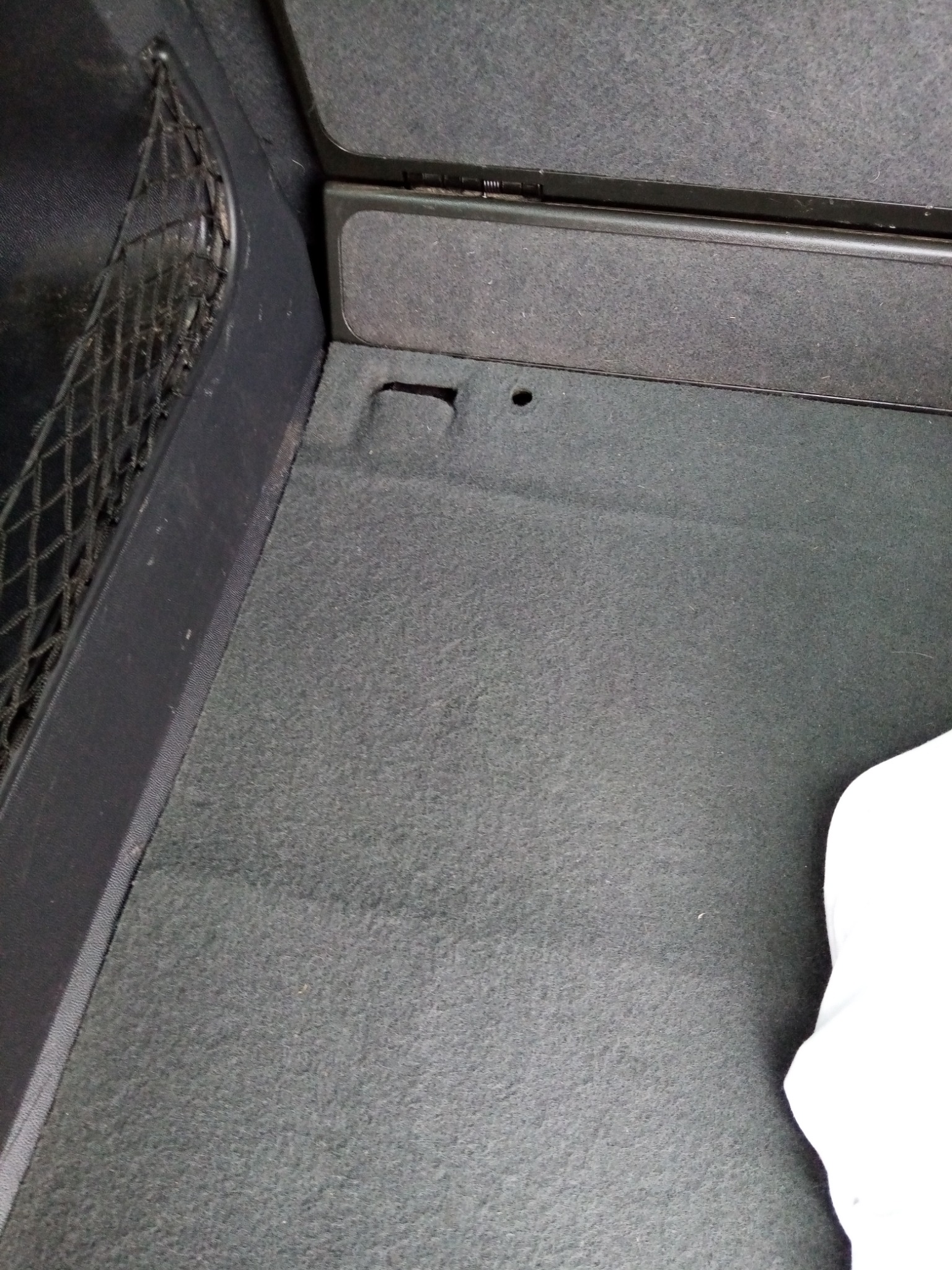 Szary dywanik w bagażniku samochodu z siatką po lewej stronie i fragmentem białej tkaniny w prawym dolnym rogu.