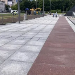 Szeroki chodnik z szarych i bordowych płyt, z widocznym fragmentem trawnika z młodą trawą, ogrodzeniem i koparką w tle.