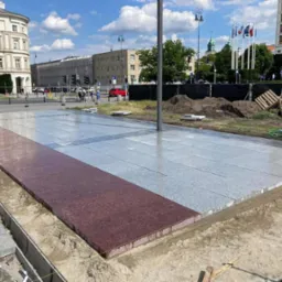 Fragment placu miejskiego z częściowo ułożoną nawierzchnią z szarej i bordowej kostki granitowej, widoczne przygotowane podłoże, w tle zabudowa miejska i maszty flagowe.