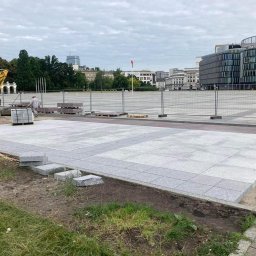 StarBRUK - Fragment placu miejskiego w trakcie budowy z wykorzystaniem szarych płyt granitowych, widoczny żółty mini-koparka i palety z materiałem, w tle budynki Warszawy.