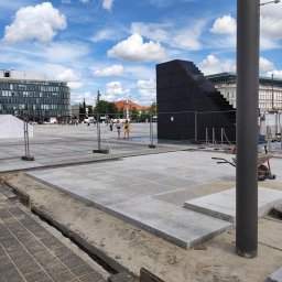 StarBRUK - Plac budowy z częściowo ułożoną szarą kostką, widoczne przygotowane podłoże, taczka i czarna konstrukcja przypominająca schody w tle, błękitne niebo z chmurami.