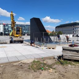 StarBRUK - Prace brukarskie na placu miejskim: żółta koparka, robotnicy układający jasnoszarą kostkę granitową wokół czarnej, schodkowej konstrukcji, w tle budynki i błękitne niebo.