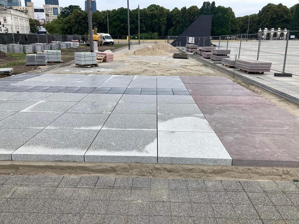 Fragment placu z ułożoną częścią nawierzchni z dużych, szarych i czerwonych płyt, palety z materiałem budowlanym w tle, teren budowy w centrum miasta.