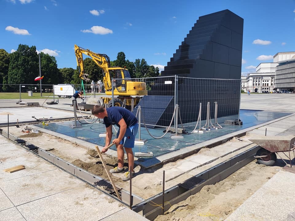 Pracownik wyrównuje piasek w wykopie pod obrzeża nowej nawierzchni placu miejskiego, w tle czarny, schodkowy monument, koparka i budynki.