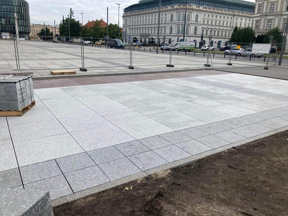 Fragment placu miejskiego z nową, jasnoszarą nawierzchnią z kamiennych płyt, widoczne palety z materiałem i ogrodzenie budowy w tle.