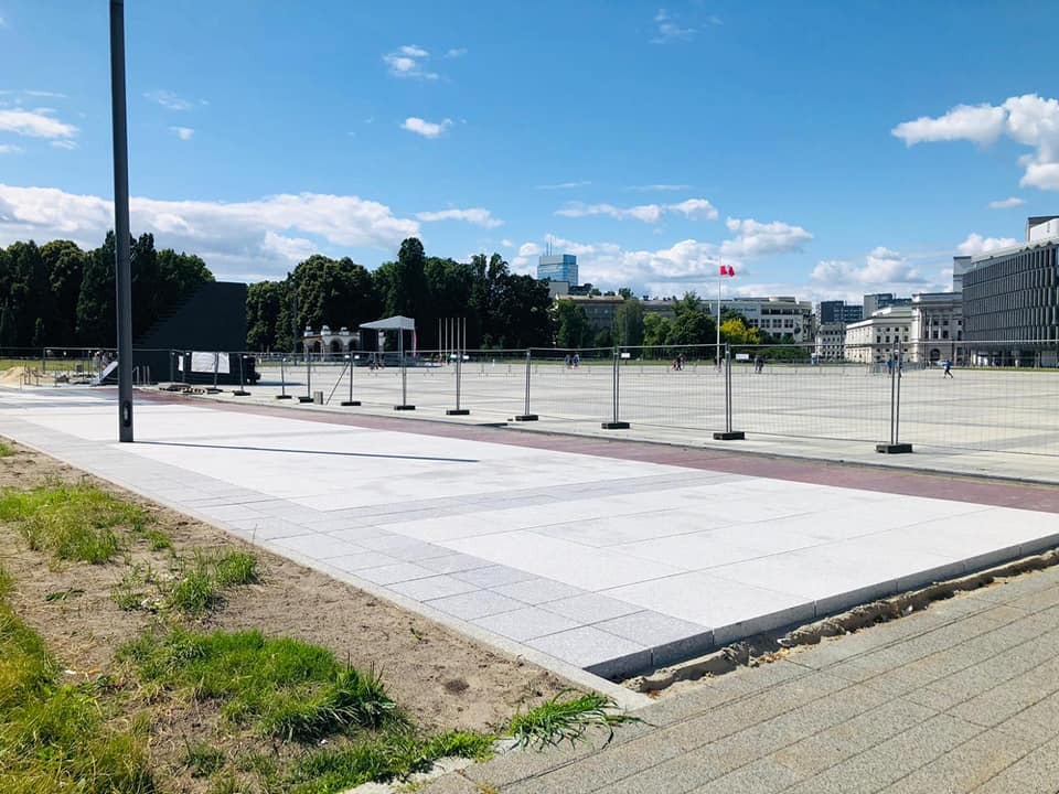 Nowa, jasnoszara nawierzchnia placu miejskiego z kostki, oddzielona od trawnika szarym obrzeżem, w tle widoczne budynki miejskie i zabezpieczenia budowy.