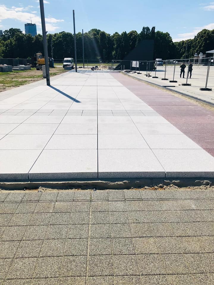 Nowa, jasna nawierzchnia z dużych płyt na placu miejskim, widoczne prace wykończeniowe i ekipa w oddali, w tle zieleń miejska i budynek.