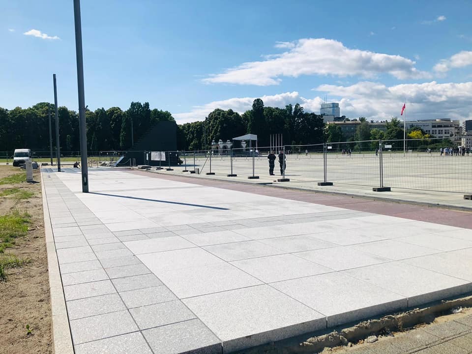 Nowo ułożona, jasnoszara nawierzchnia z kwadratowych płyt, widok z perspektywy chodnika, z widocznymi słupami oświetleniowymi, ogrodzeniem tymczasowym i budynkami w tle.