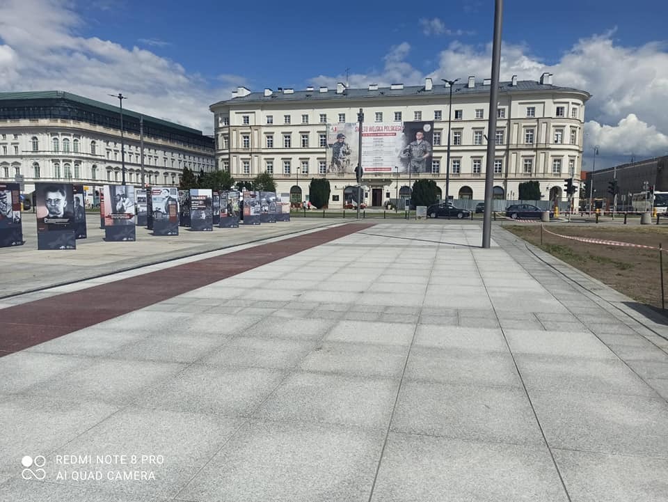 Szara kostka brukowa na placu miejskim z ekspozycją plenerową, w tle budynki z banerami i pochmurne niebo.