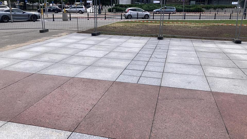 Szara i czerwona kostka ułożona w regularny wzór na placu miejskim, tymczasowe barierki ochronne w tle.
