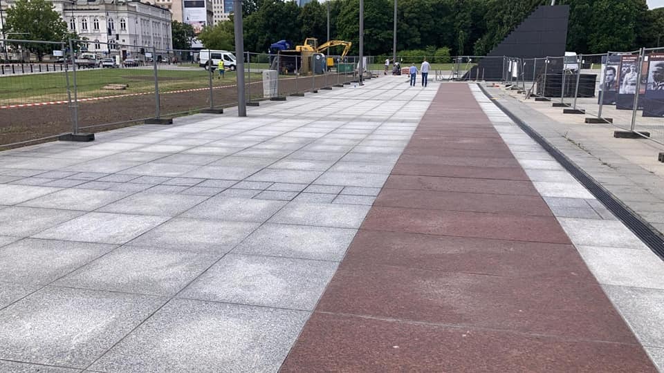 Szeroki chodnik z szarych i bordowych płyt, z widocznym fragmentem trawnika z młodą trawą, ogrodzeniem i koparką w tle.