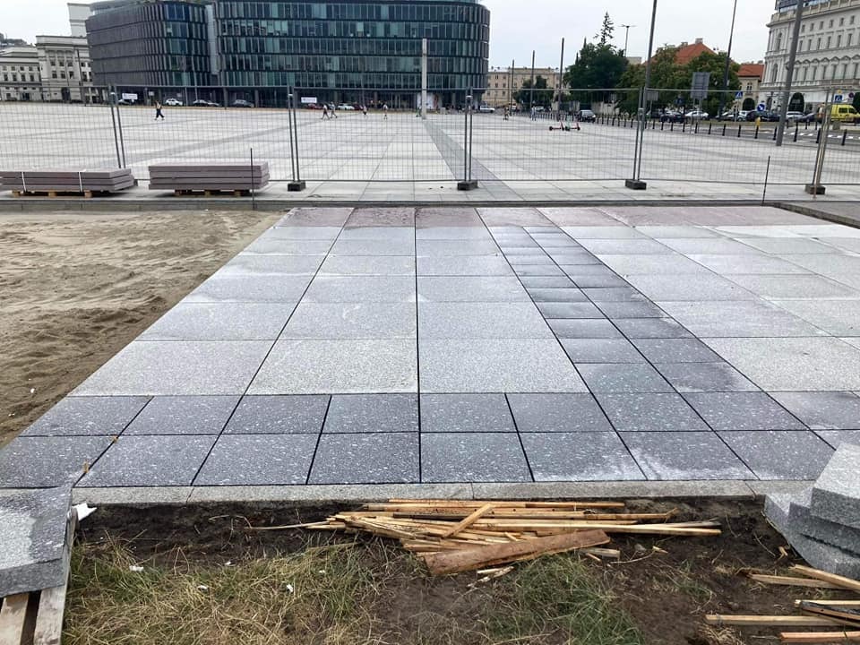 Układanie szarej kostki brukowej na dużej powierzchni placu miejskiego, widoczne drewniane kliny i piasek przygotowany pod kolejne rzędy, w tle budynki administracyjne i barierki zabezpieczające...