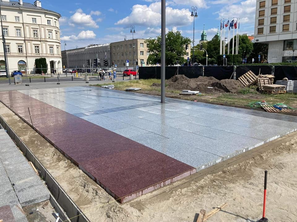 Fragment placu miejskiego z częściowo ułożoną nawierzchnią z szarej i bordowej kostki granitowej, widoczne przygotowane podłoże, w tle zabudowa miejska i maszty flagowe.