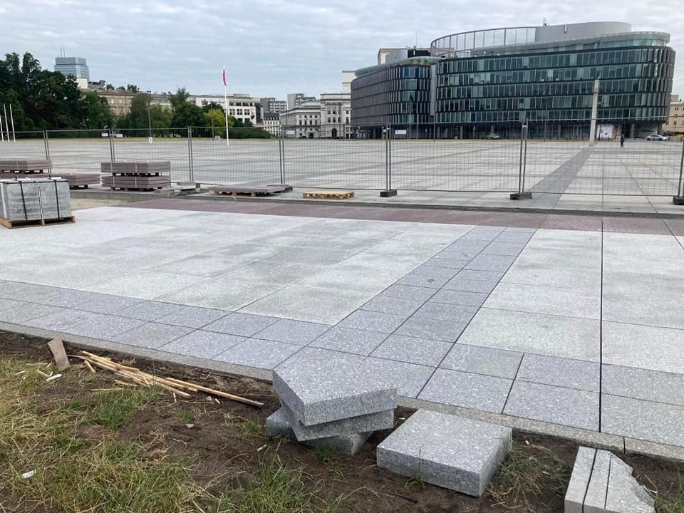 Fragment placu miejskiego z nową, szarą nawierzchnią z kostki, widoczne palety z materiałem i budynki w tle.