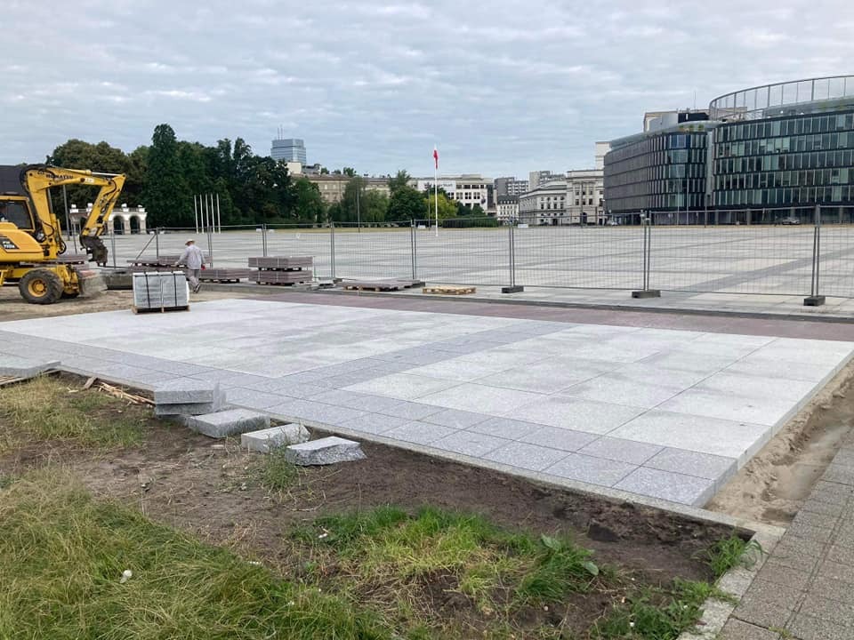 Fragment placu miejskiego w trakcie budowy z wykorzystaniem szarych płyt granitowych, widoczny żółty mini-koparka i palety z materiałem, w tle budynki Warszawy.