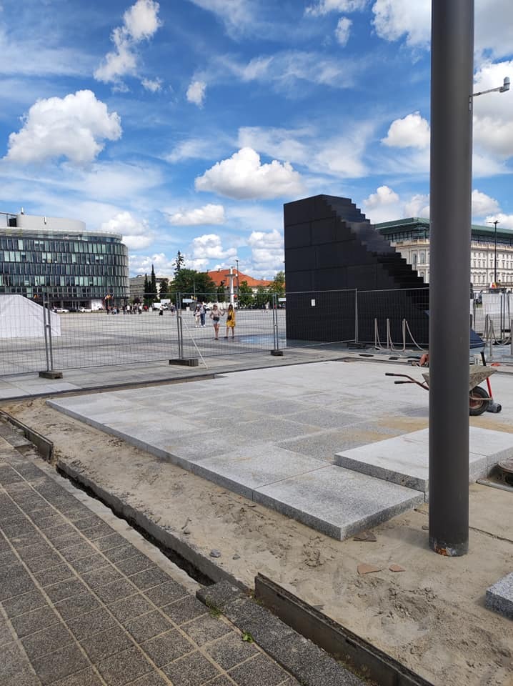 Plac budowy z częściowo ułożoną szarą kostką, widoczne przygotowane podłoże, taczka i czarna konstrukcja przypominająca schody w tle, błękitne niebo z chmurami.