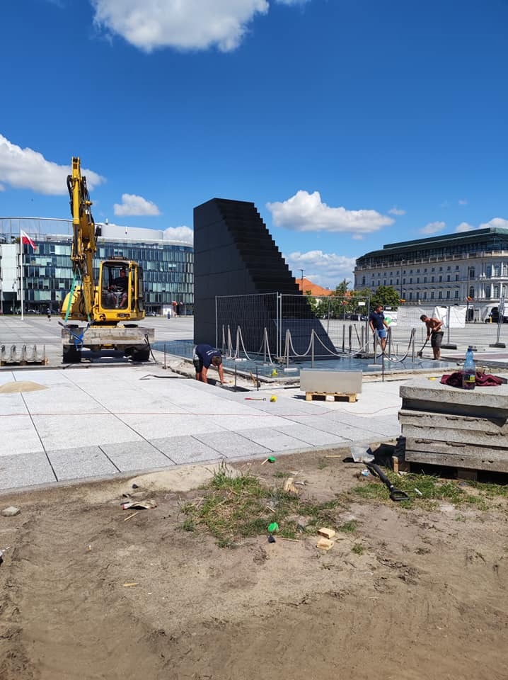 Prace brukarskie na placu miejskim: żółta koparka, robotnicy układający jasnoszarą kostkę granitową wokół czarnej, schodkowej konstrukcji, w tle budynki i błękitne niebo.