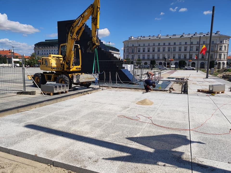 Układanie szarej kostki brukowej na placu miejskim, żółta koparka podnosi element konstrukcyjny, widoczny pracownik i piasek.