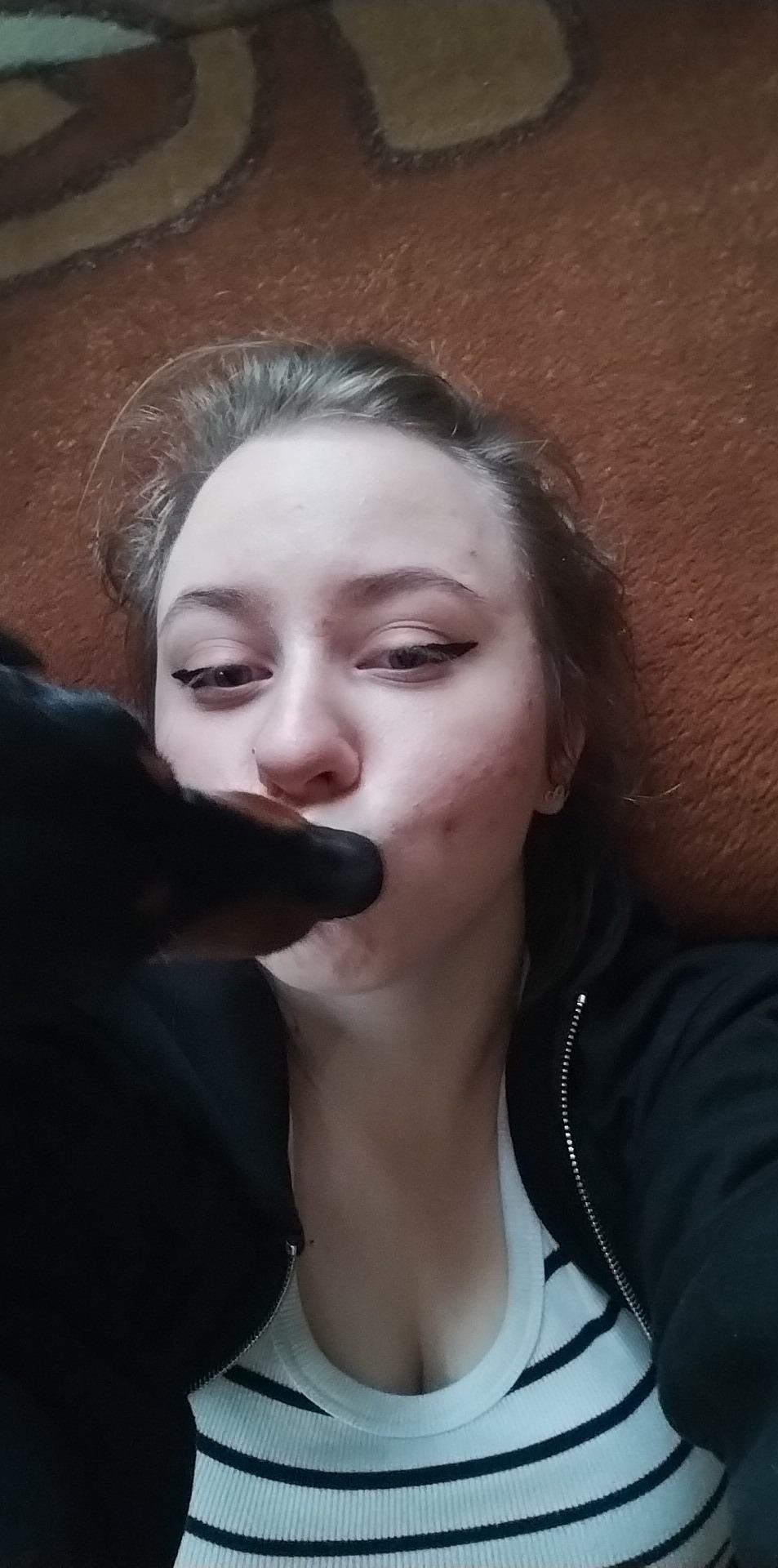 Młoda kobieta z eyelinerem leżąca na brązowym dywanie, pies rasy jamnik liże ją w policzek.