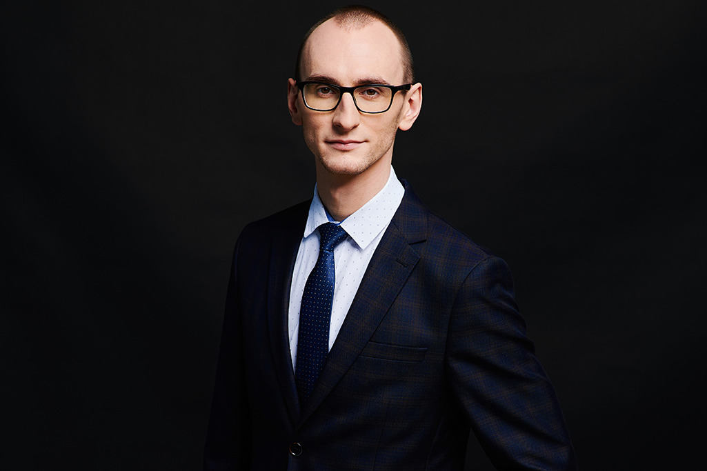 Portret mężczyzny w okularach i granatowym garniturze w kratę na ciemnym tle, z subtelnym uśmiechem i spojrzeniem w obiektyw.