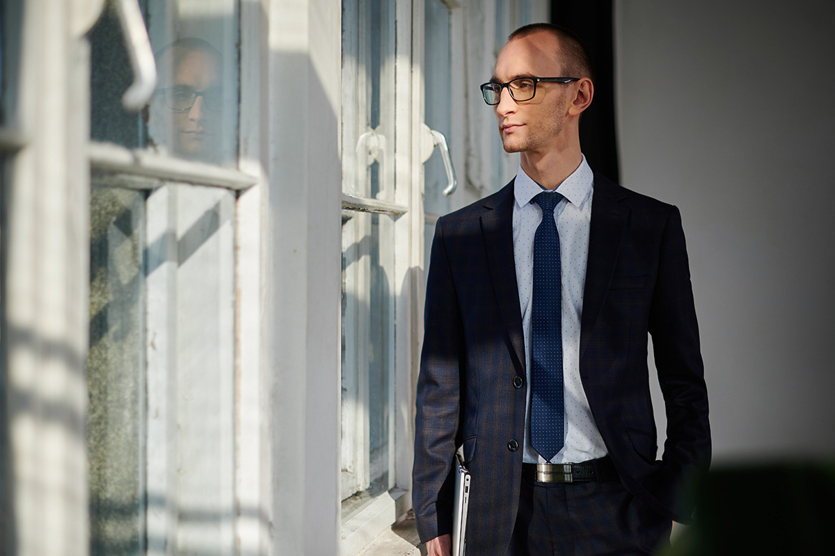 Mężczyzna w garniturze, z laptopem pod pachą, stoi przy oknie z odbiciem, spoglądając w bok.