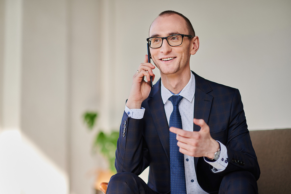 Uśmiechnięty mężczyzna w garniturze i okularach rozmawia przez telefon, gestykulując dłonią. Widoczny zegarek na nadgarstku i pierścionek na palcu.