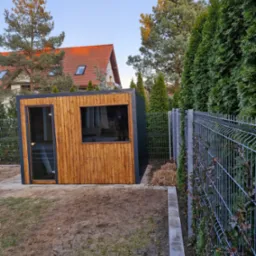 Nowoczesna, drewniana konstrukcja z dużym oknem i przeszklonymi drzwiami, prawdopodobnie altana lub domek ogrodowy, umieszczona na działce z widocznym ogrodzeniem z siatki i zielenią w tle.