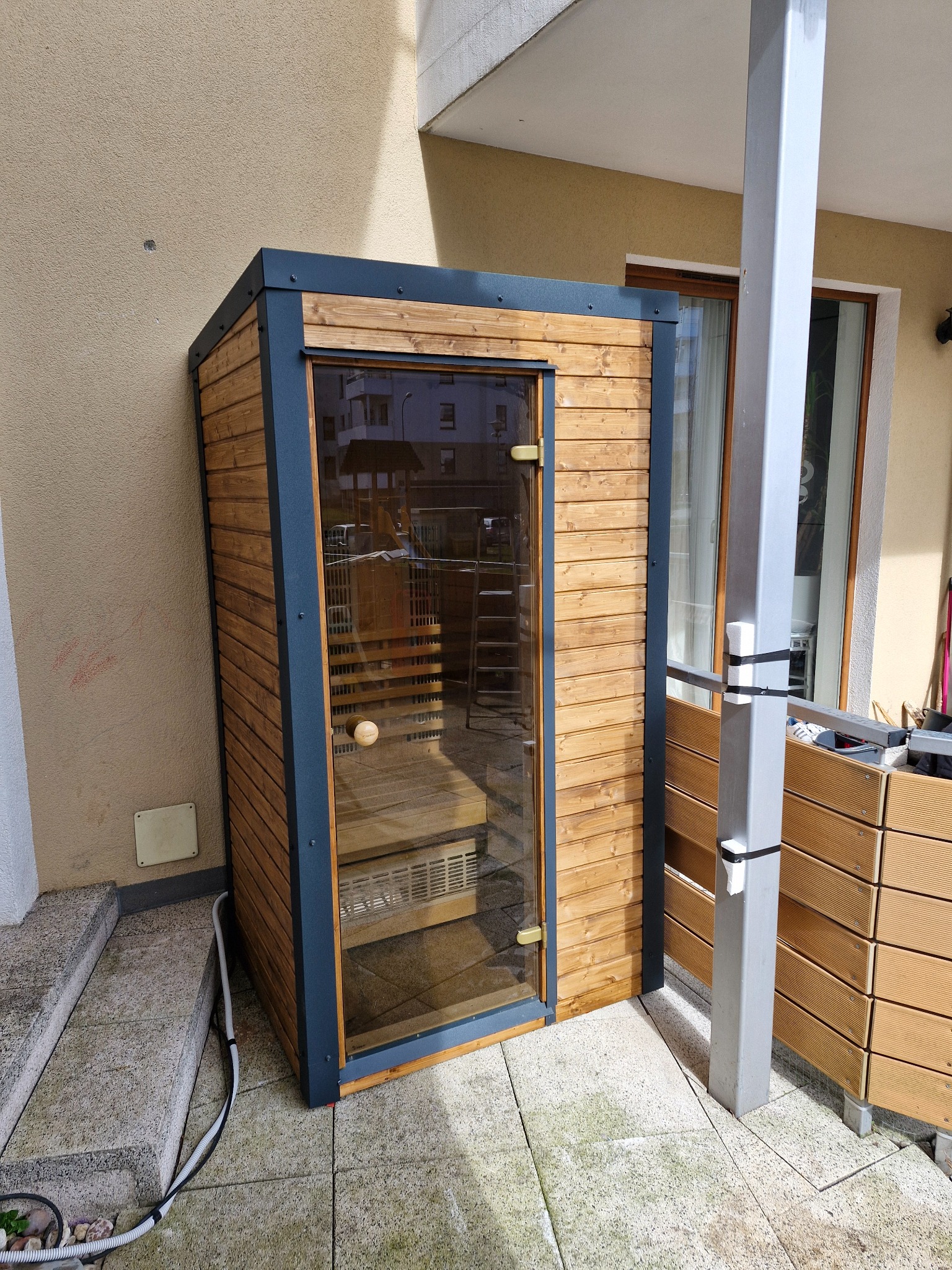 Drewniana sauna zewnętrzna z przeszklonymi drzwiami, umieszczona na tarasie obok budynku. Widoczne schody wejściowe i elementy instalacji elektrycznej.