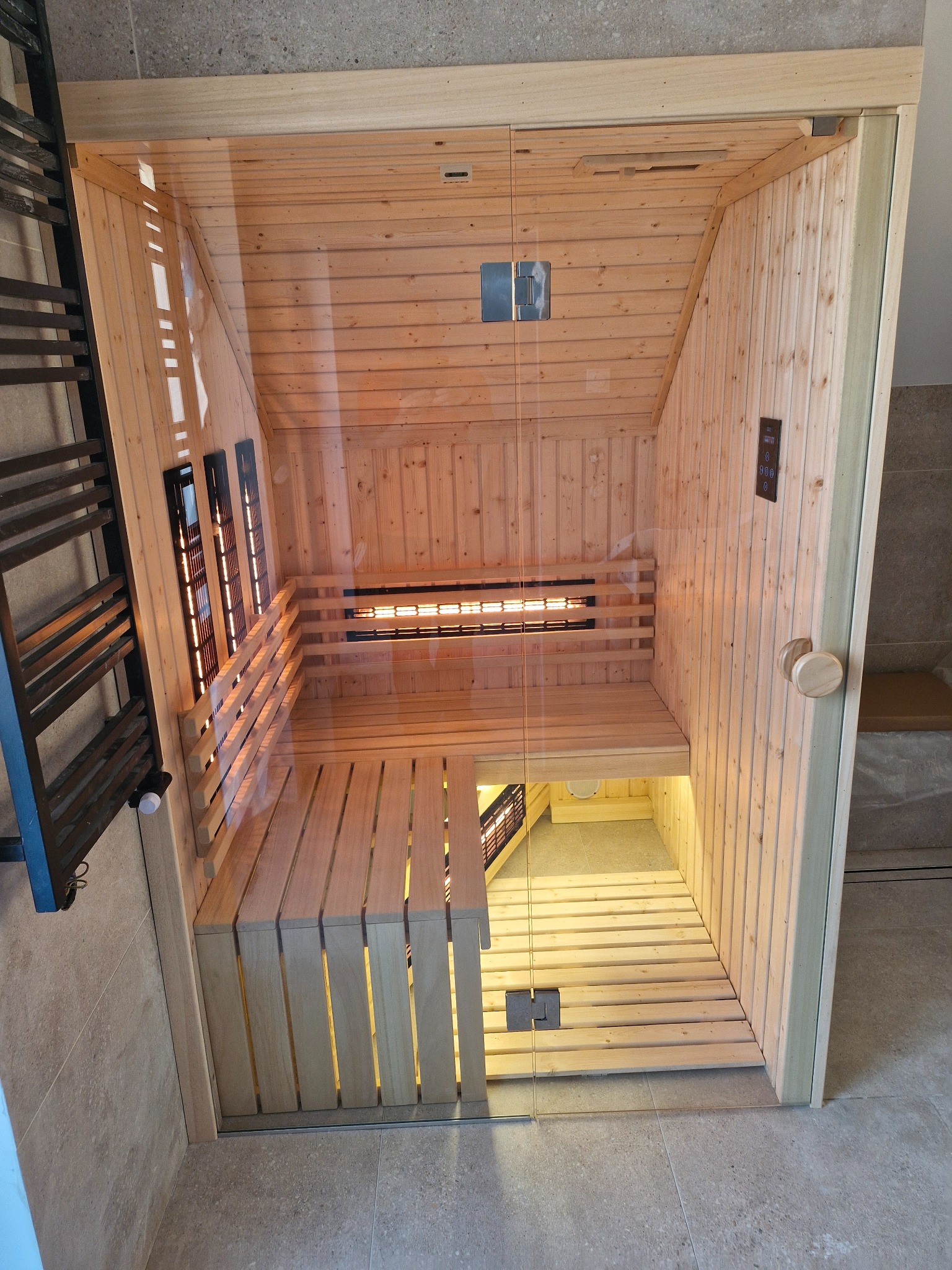 Wnętrze nowoczesnej, drewnianej kabiny z przeszklonymi drzwiami, z widocznymi panelami grzewczymi i oświetleniem, ustawionej w łazience z beżowymi płytkami.