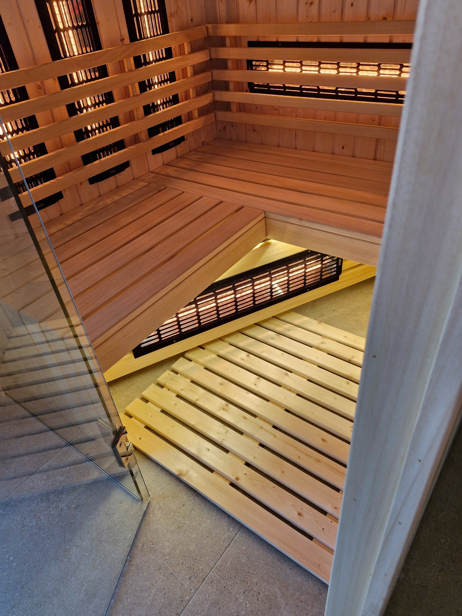 Wnętrze sauny infrared z drewnianymi ławkami i panelami grzewczymi, widok z perspektywy wejścia z przeszklonymi drzwiami w Łochocinie.
