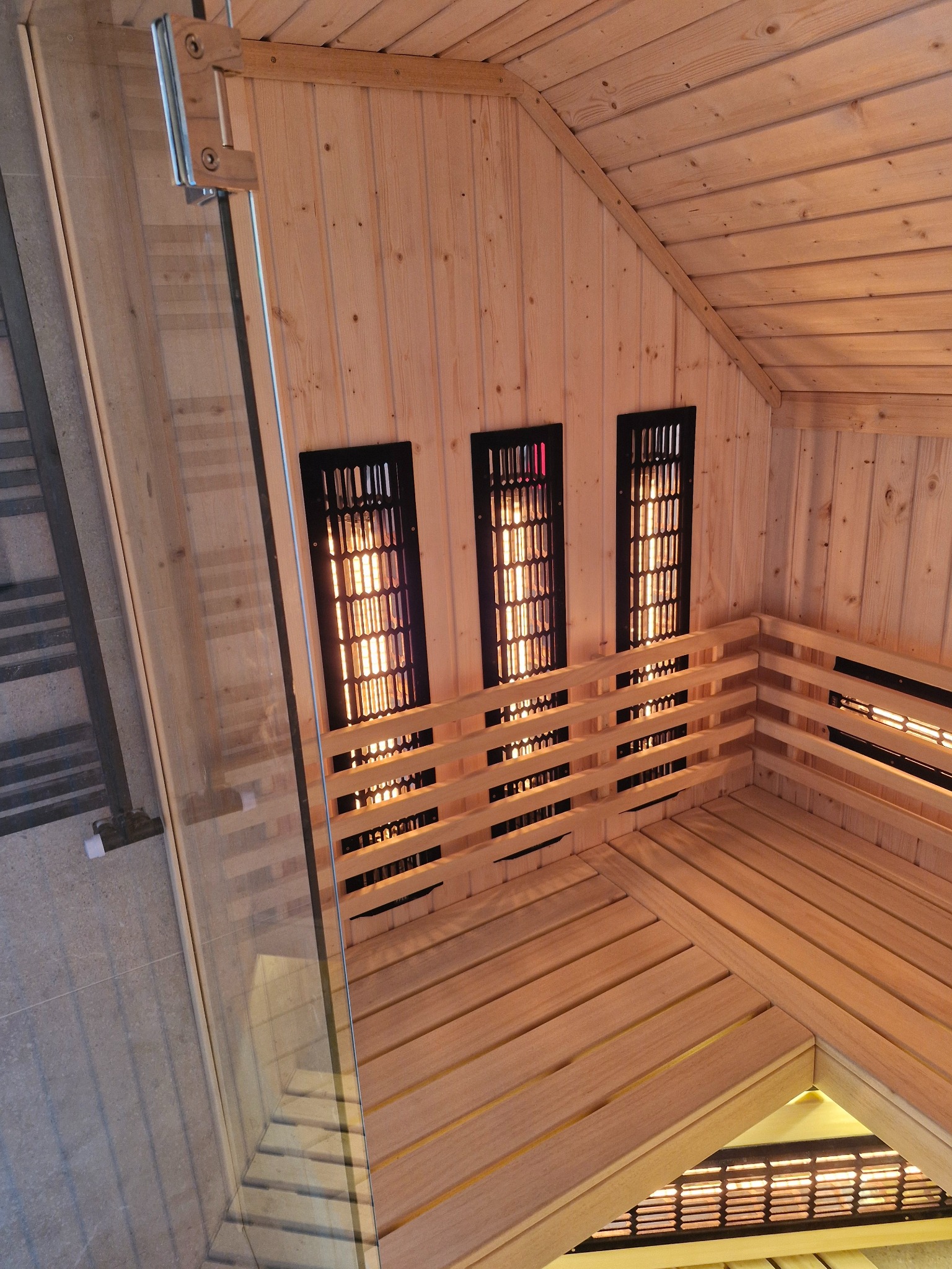 Wnętrze sauny z drewnianymi ławkami, trzema pionowymi panelami grzewczymi w czarnych obudowach i szklaną ścianą z metalowymi zawiasami.
