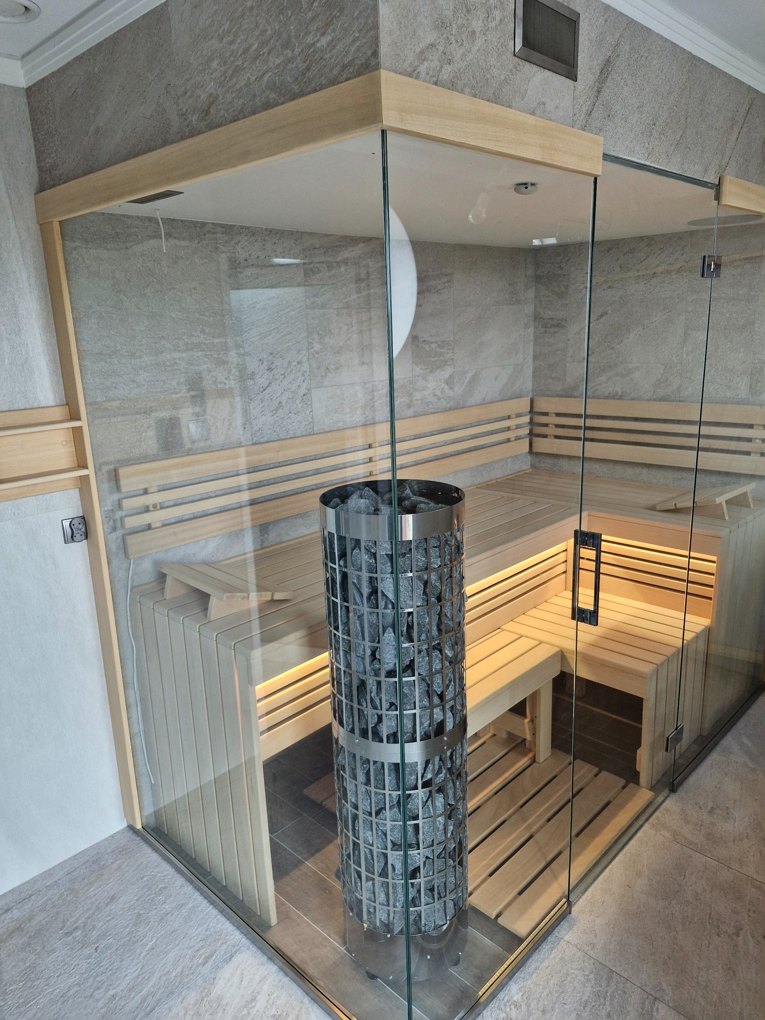 Szklana kabina z drewnianymi ławkami wewnątrz, wypełniona kamieniami grzewczymi w metalowej obudowie, podświetlona od dołu.