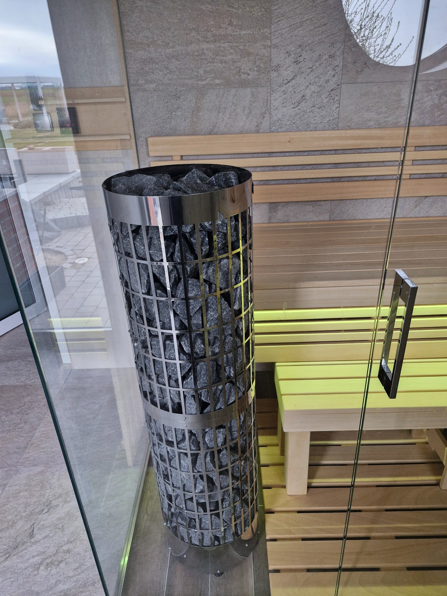 Wnętrze nowoczesnej kabiny z przeszklonymi ścianami, z piecem na kamienie w metalowej obudowie siatkowej oraz drewnianymi ławkami podświetlonymi od dołu.