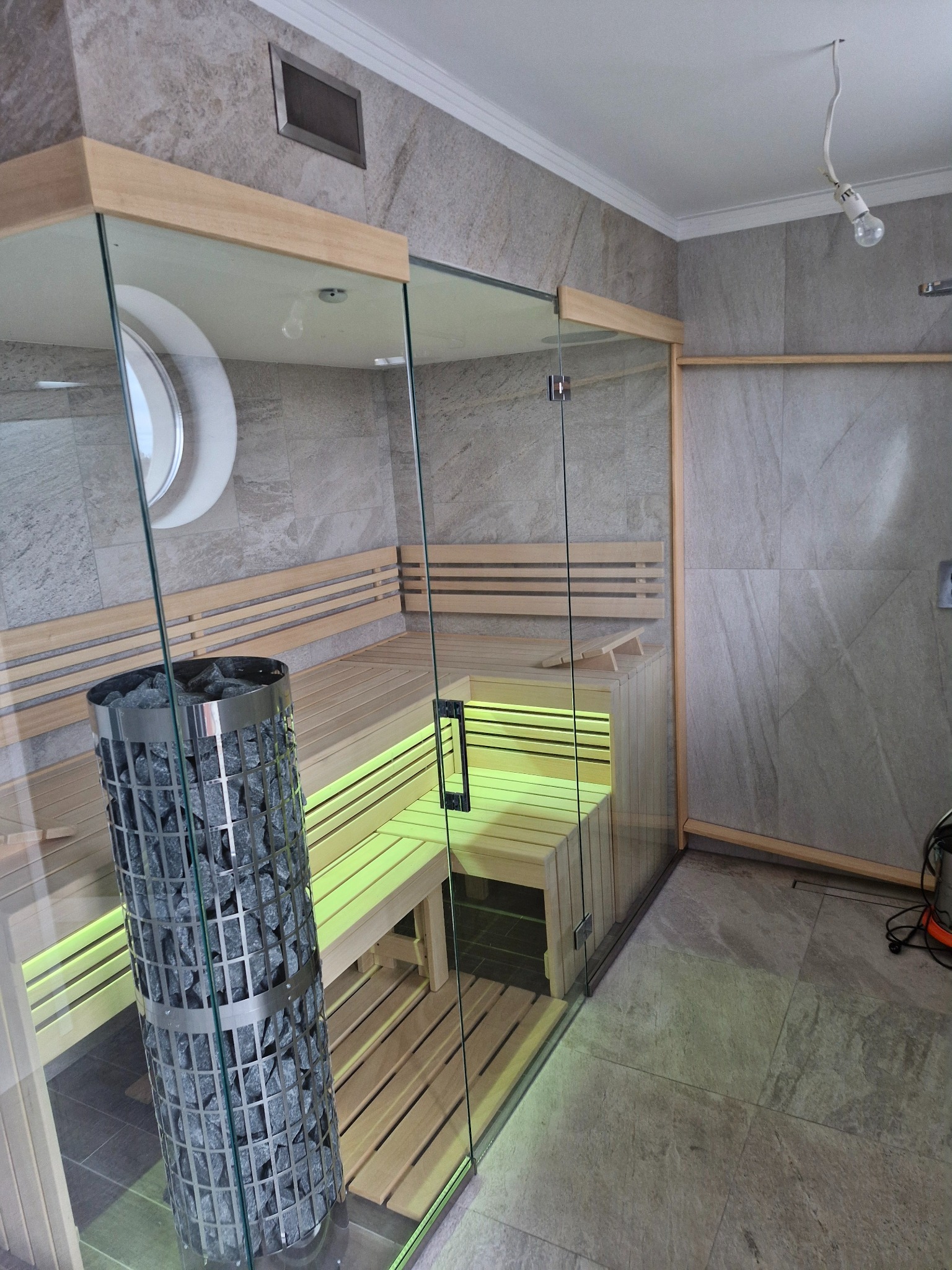 Nowoczesna kabina z drewna i szkła z piecem na kamienie, oświetlona od spodu na szaro-beżowym tle płytek.