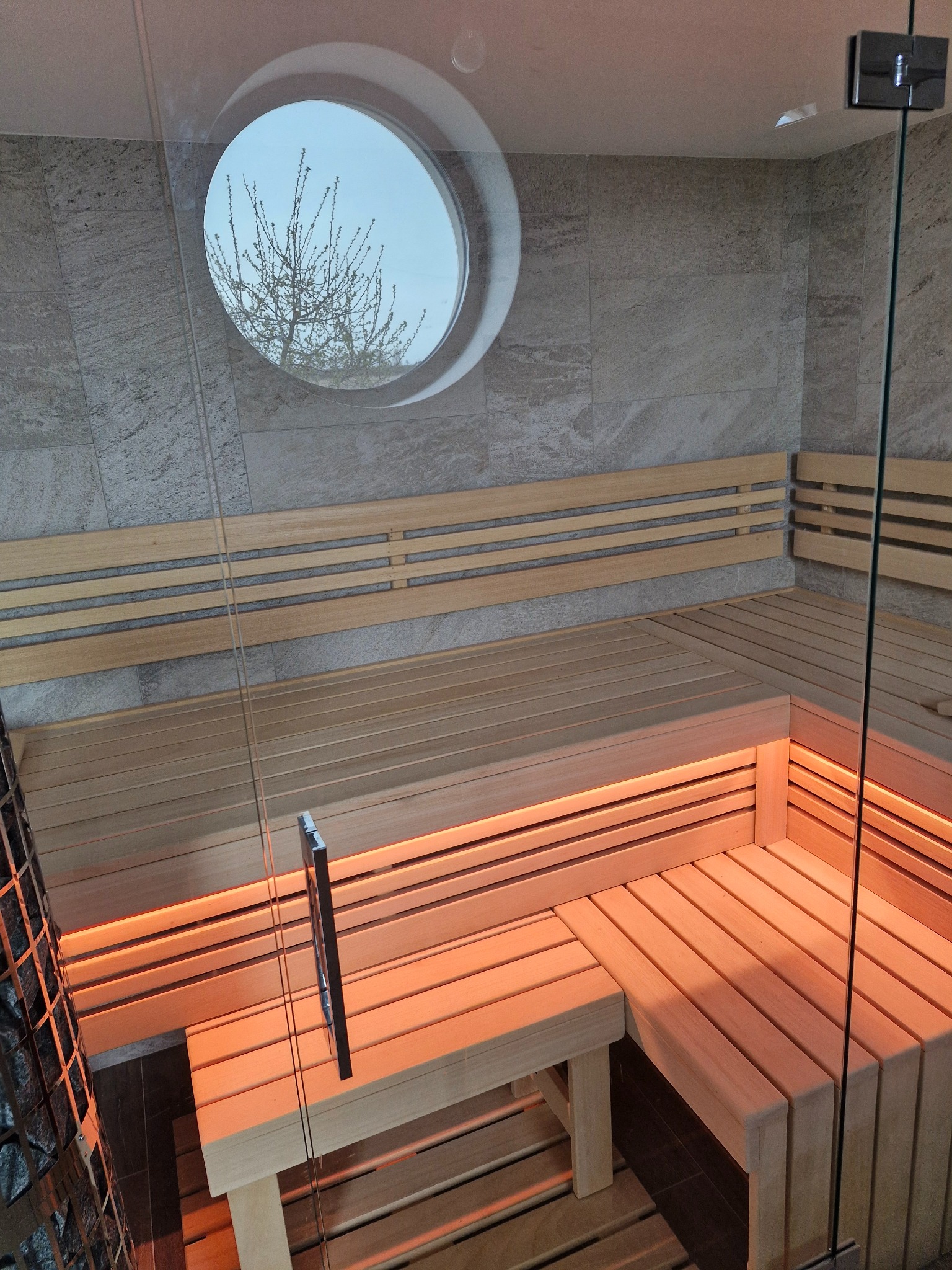 Wnętrze nowoczesnej kabiny z jasnego drewna z oknem w kształcie koła, przez które widać drzewo, podświetlone pomarańczowym światłem, z elementami kamienia na ścianach.