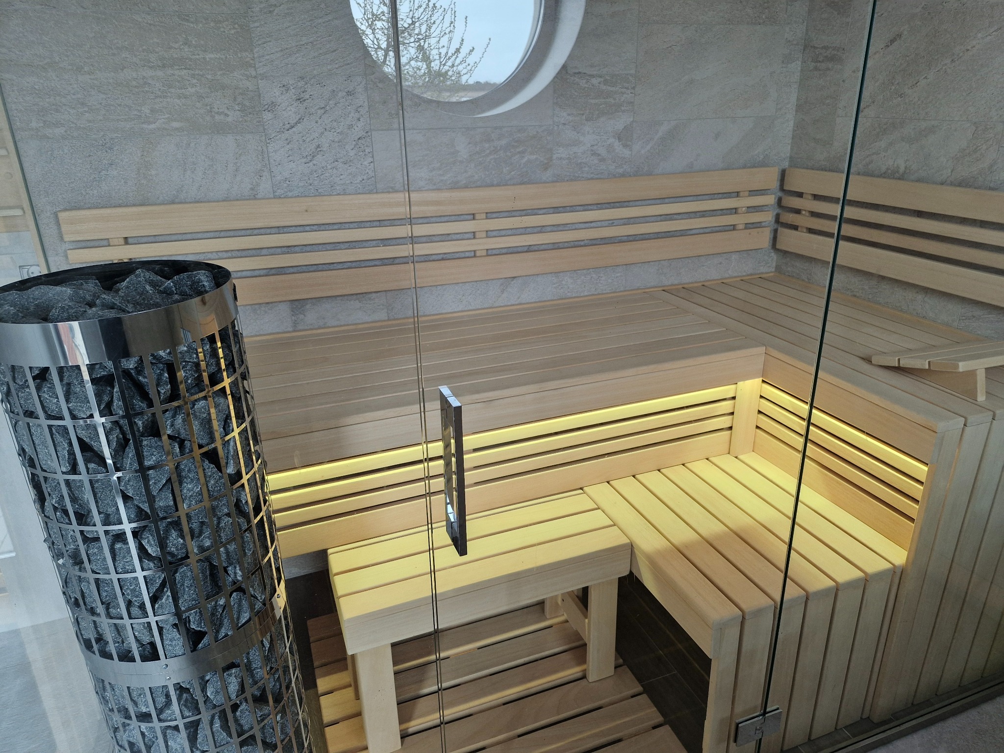 Nowoczesna kabina z drewna w jasnych barwach z oświetleniem LED pod ławkami, piec na kamienie w metalowej obudowie, okrągłe okno w górnej części ściany.
