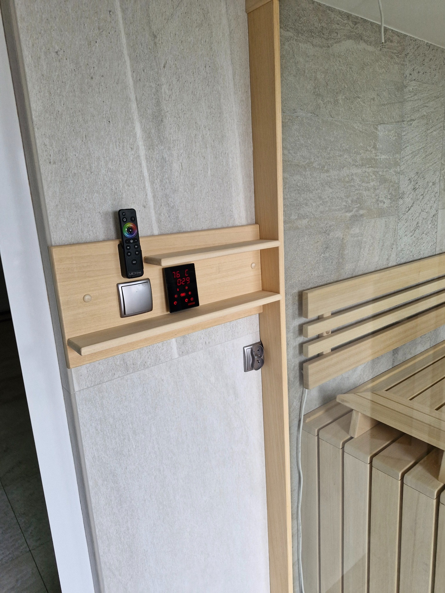 Wnętrze sauny: drewniane ławki, panel sterowania z wyświetlaczem temperatury i pilot do oświetlenia, jasne płytki na ścianach.