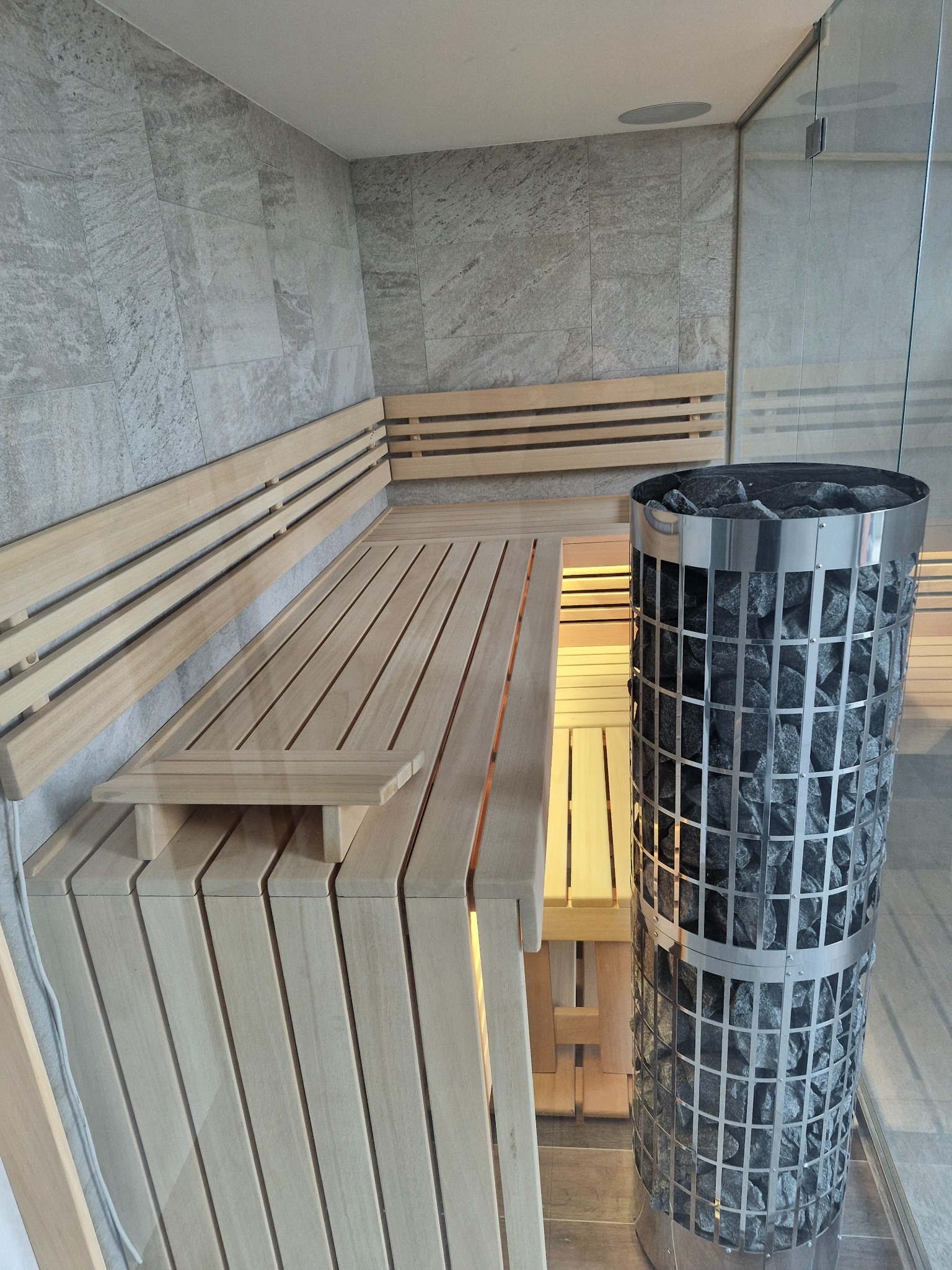 Wnętrze wykończonej kabiny z jasnego drewna z piecem na kamienie w metalowej obudowie, widoczne szare płytki na ścianach i szklane drzwi.