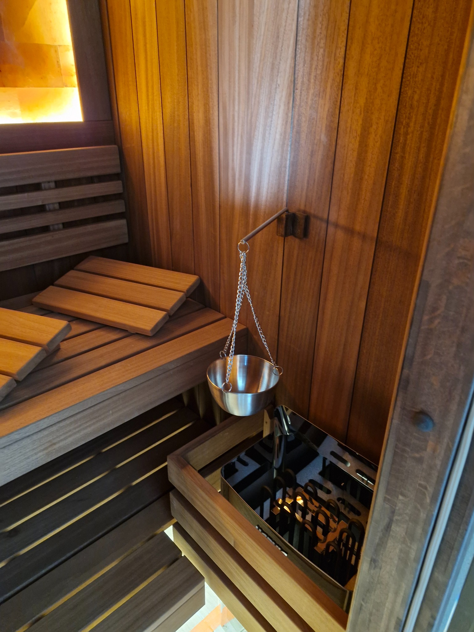 Drewniana sauna z oświetleniem solnym, ławkami i metalowym naczyniem na łańcuszku nad piecem. Widoczne pionowe deski boazerii i elementy wyposażenia.