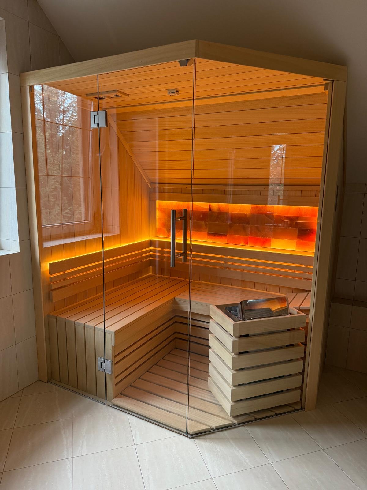 Nowoczesna sauna narożna z przeszklonymi ścianami, oświetleniem LED i panelem solnym, umieszczona w pomieszczeniu z jasnymi płytkami podłogowymi.